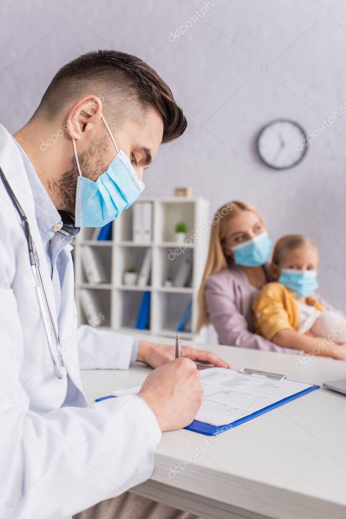 Doctor in medical mask writing on clipboard near blurred mother and daughter in hospital 