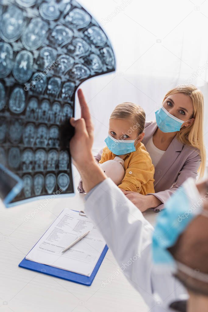 Mother and kid in medical masks looking at pediatrician with mri scan on blurred foreground 