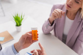 Teilansicht des Arztes zeigt auf Behälter mit Pillen nahe verschwommener Frau