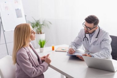 Doktor, sarışın kadın, dizüstü bilgisayar ve haplı konteynır yanında reçete yazıyor.