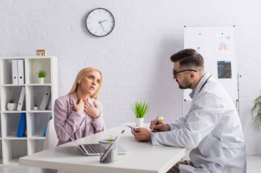 Doktor, muayene sırasında boğazı ağrıyan bir kadına bakarken panoya yazı yazıyor.