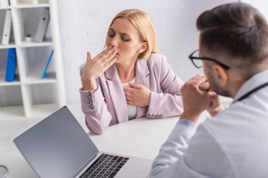 Kadın, muayene odasında boş ekranlı bulanık bir doktor ve dizüstü bilgisayarın yanında öksürüyor.