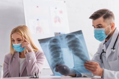 Doktor, tıbbi maskeli hasta bir kadının yakınındaki röntgene bakıyor.