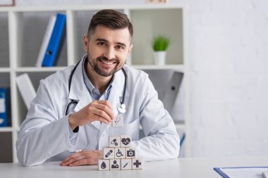 Masasında tıbbi ikonlar olan küp piramidinin yanında gülümseyen beyaz önlüklü bir doktor.