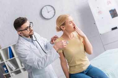 Hastalıklı kadın hastanede teşhis sırasında doktorun yanında öksürüyor.