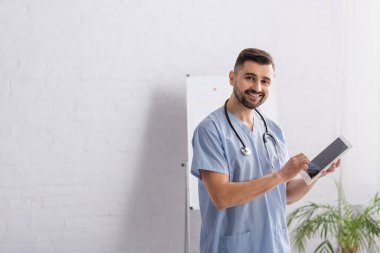 Mavi üniformalı doktor elinde dijital tablet tutarken kameraya gülümsüyor.