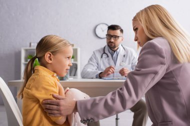 Hastanede bulanık bir doktorun yanında annesine bakan endişeli bir çocuk.