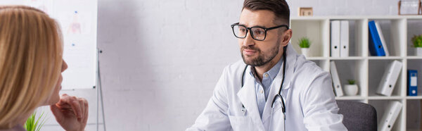 doctor in white coat and eyeglasses near blonde patient in clinic, banner