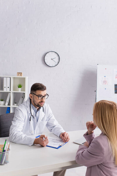 doctor in white coat talking to blonde woman near clipboard with prescription