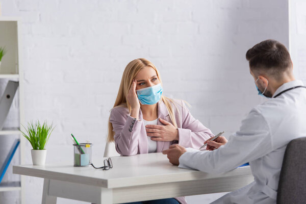 blonde woman in protective mask suffering from headache near physician