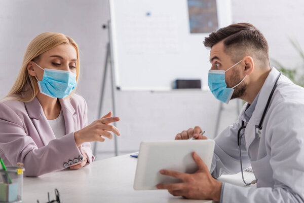 doctor and patient in medical masks pointing at digital tablet during appointment