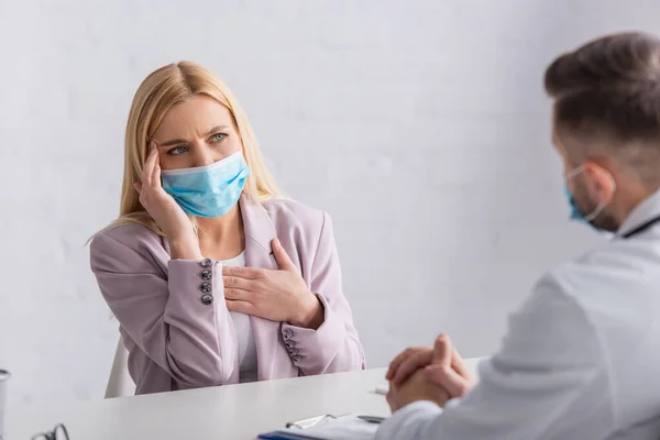 Mujer Enferma Con Máscara Médica Tocando Cabeza Pecho Durante Consulta —  Fotos de Stock