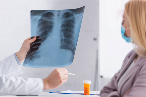 обрезанный вид врача, указывающего ручкой на контейнер с таблетками рядом с флюорографией сканирования и пациента в медицинской маске