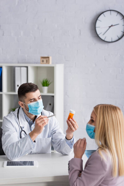 doctor in medical mask pointing at pills near blonde woman