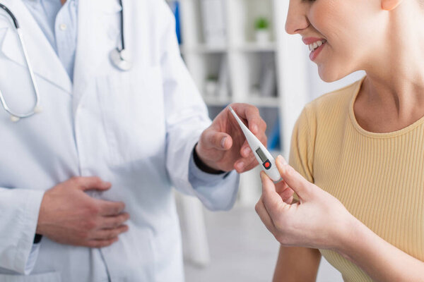 частичный взгляд улыбающейся женщины, забирающей электронный термометр у врача в клинике