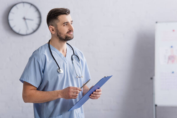 smiling doctor in blue uniform looking away while holding clipboard