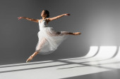 Boční pohled na balerína v bílých šatech skákání na slunci na šedém pozadí 