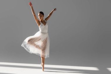 Brunette ballerina in white skirt dancing with closed eyes on grey background with sunlight clipart