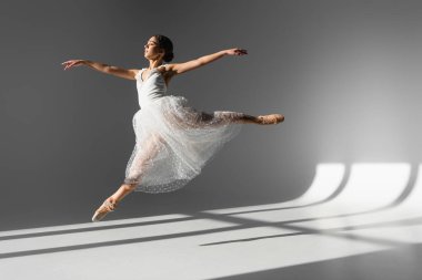 Side view of ballerina in white dress jumping in sunlight on grey background  clipart