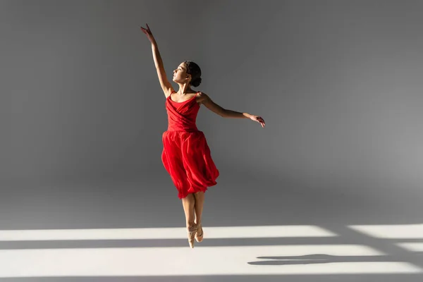 Zijaanzicht Van Ballerina Rode Jurk Springen Grijze Achtergrond Met Zonlicht — Stockfoto