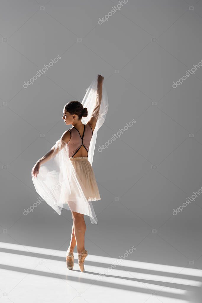 Side view of elegant ballerina dancing with white cloth on grey background with sunlight