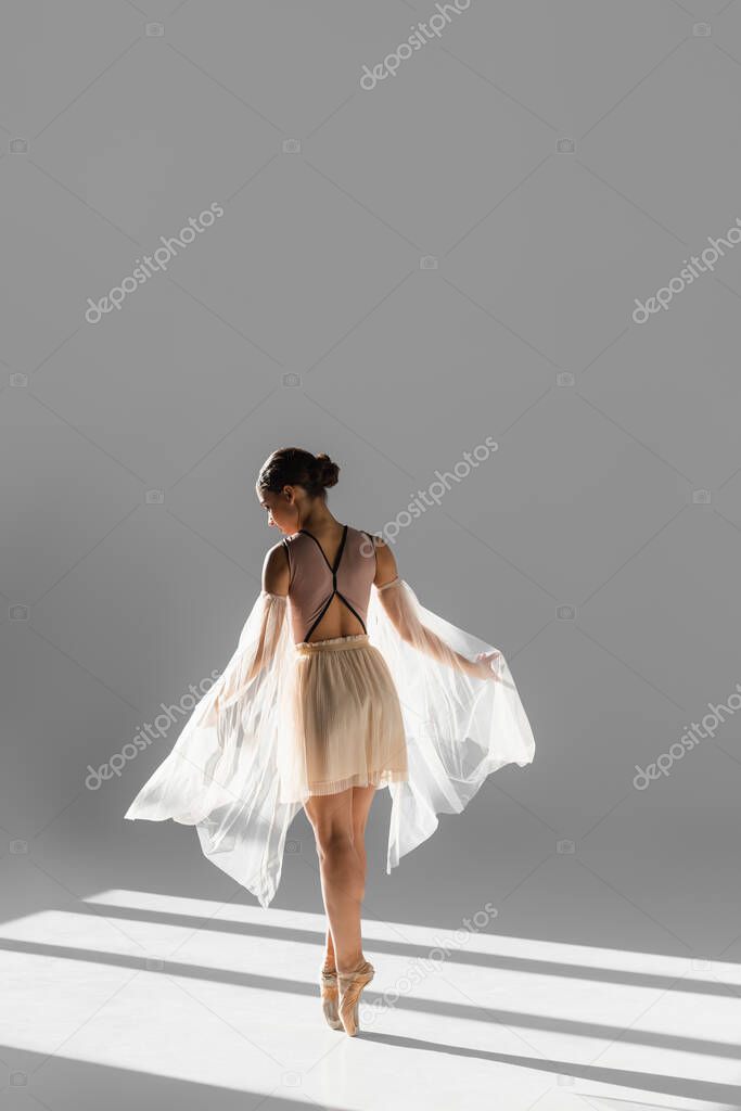 Side view of ballerina in pointe shoes dancing with cloth in sunlight on grey background 