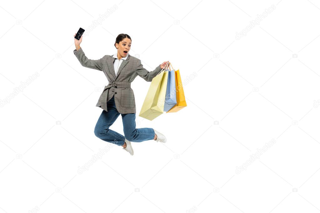 Amazed woman with smartphone holding shopping bags and jumping isolated on white 