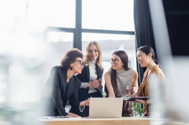 Gülümseyen Afrikalı Amerikalı iş kadını, çok ırklı iş arkadaşlarıyla konuşurken dizüstü bilgisayarını işaret ediyor.