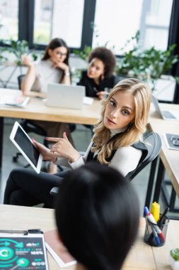 Ofisteki bulanık iş ortağının yanındaki dijital tableti gösteren sarışın kadın.