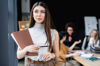 Esmer iş kadını kağıt klasörü ve kalemle çok ırklı iş arkadaşlarının yanında duruyor.