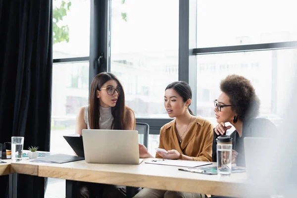 Empresarias Multiétnicas Hablando Cerca Computadora Portátil Oficina Primer Plano Borrosa — Foto de Stock