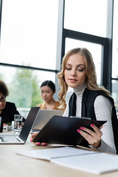 Blondynka Bizneswoman Patrząc Schowek Podczas Pracy Pobliżu Laptopa Zamazane Międzyrasowych — Zdjęcie stockowe