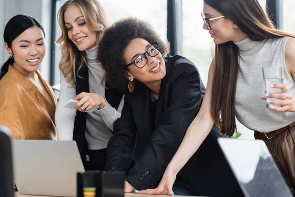 Африканская американская бизнесвумен улыбается рядом с ноутбуком и многонациональными деловыми партнерами