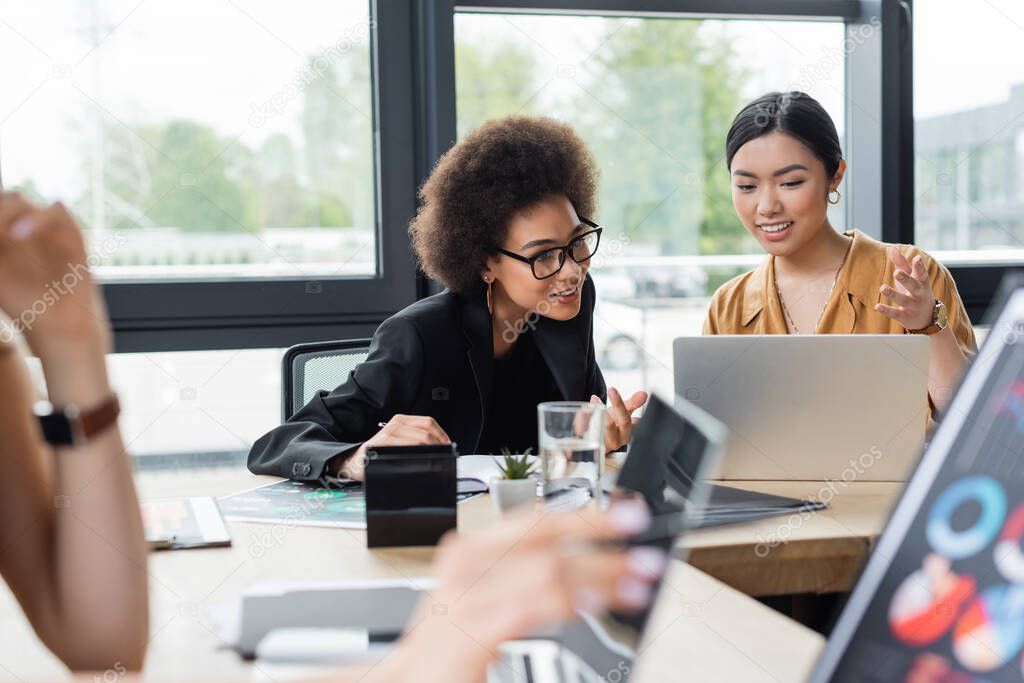 positive asian businesswoman pointing at laptop while working with african american colleague