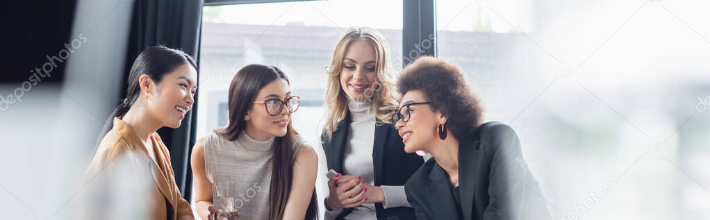 cheerful multicultural businesswomen talking in office on blurred foreground, banner