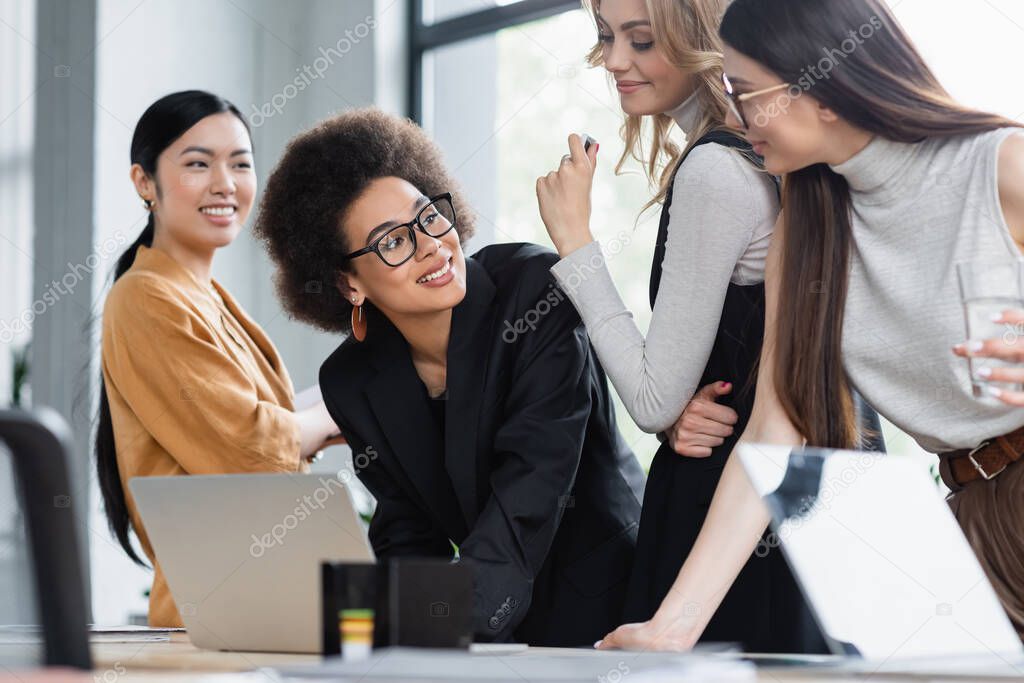 african american businesswoman smiling near multiethnic colleagues near laptop in office