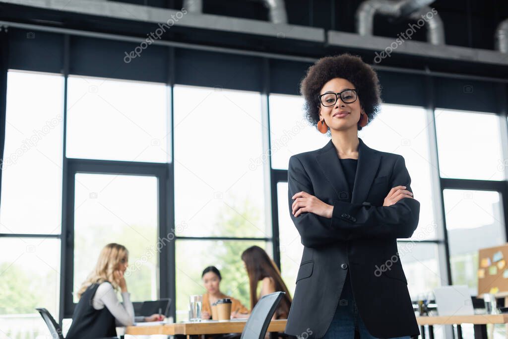happy african american businesswoman looking at camera while standing with crossed arms in office