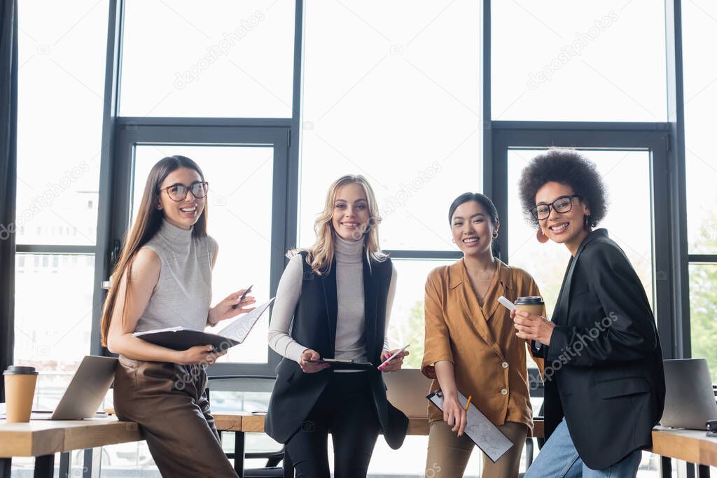 young and happy multiethnic businesswomen smiling at camera near window in office