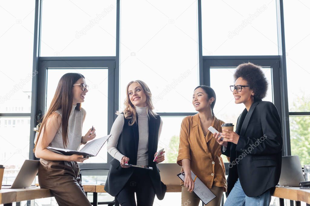 cheerful multiethnic businesswomen with documents talking during coffee break