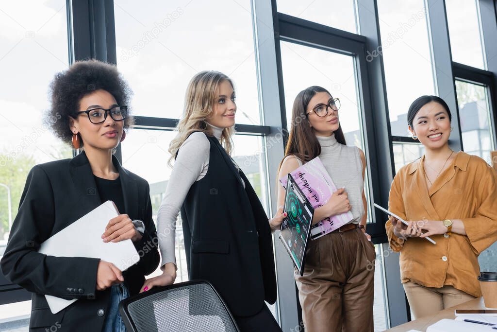 smiling multicultural businesswomen looking away while standing in office