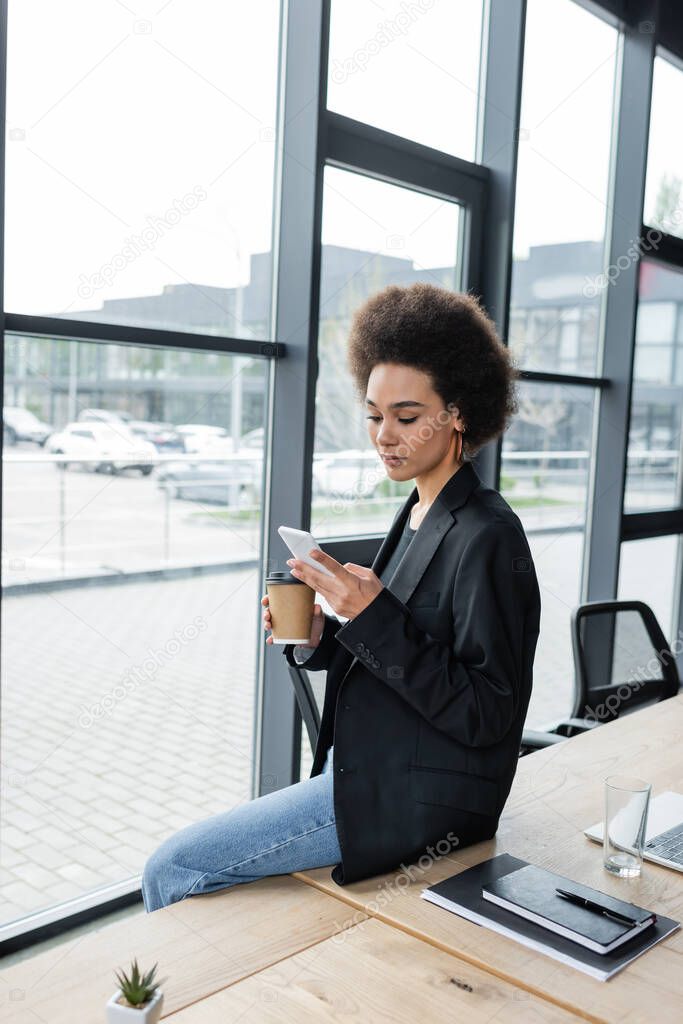 stylish african american businesswoman sitting on desk with coffee and messaging on smartphone