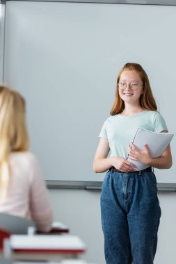 Smiling schoolgirl holding notebook near erase board and classmate  clipart