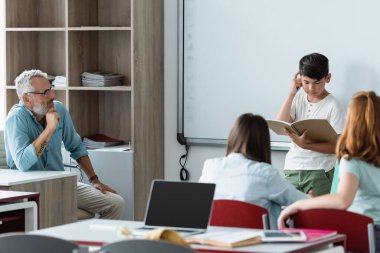 Asyalı okul çocuğu silme tahtası ve öğretmenin yanındaki deftere bakıyor. 