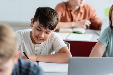 Gülümseyen Asyalı okul çocuğu sınıfta deftere yazı yazıyor. 