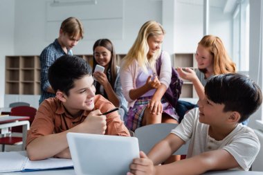 Cheerful asian child holding digital tablet near friend and classmates  clipart