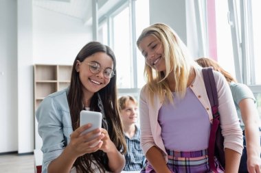 Positive teen schoolkids using cellphone in classroom  clipart