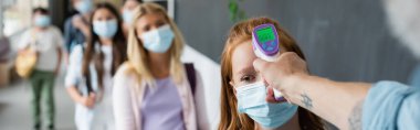 Tıp maskeli kız öğrencinin derecesini ölçen öğretmenin elinde pirometre, afiş
