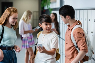 Okulun koridorunda arkadaşlarının yanında akıllı telefon tutarken gülümseyen Asyalı çocuk.