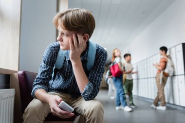 depressed boy with smartphone sitting alone in school corridor near teenagers on blurred background clipart