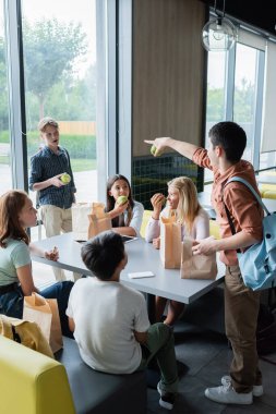 teenage girl pointing at classmate near pupils having lunch in dining room clipart